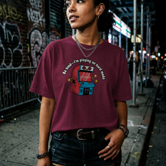 Translation missing: en.Maroon shirt with text Be kind—I'm playing on hard mode above a cracked retro arcade cabinet with Game Over