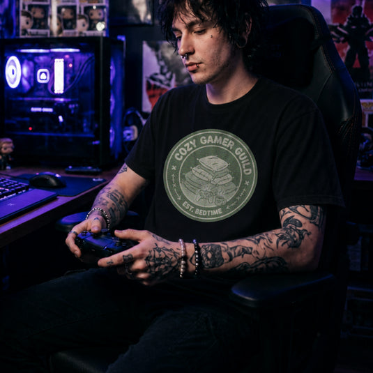 Translation missing: en.Black t-shirt with a green badge showing stacked pillows, a controller, and text Cozy Gamer Guild Est. Bedtime