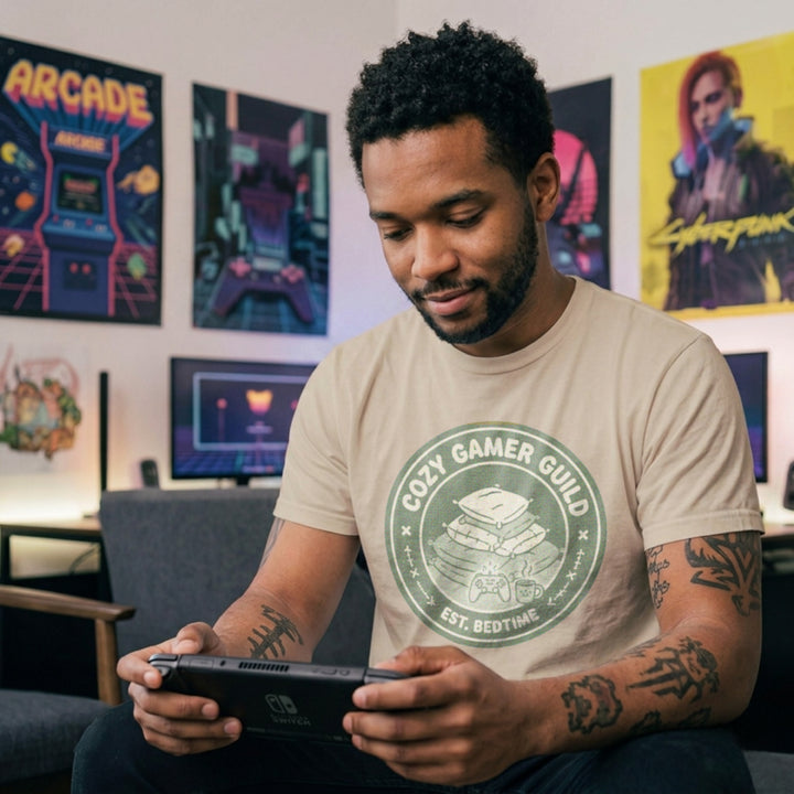 Translation missing: en.Sand t-shirt with a green badge showing stacked pillows, a controller, and text Cozy Gamer Guild Est. Bedtime