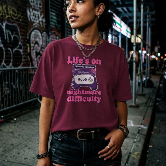 Translation missing: en.Maroon tee with pink text reading Life's on nightmare difficulty next to a sad controller and difficulty selector
