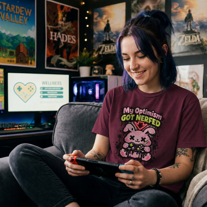 Translation missing: en. Maroon t-shirt with a pink cartoon character and text, My Optimism Got Nerfed in a gaming-themed design