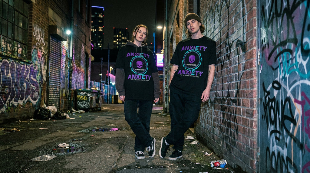 Two young men standing in a graffiti-covered urban alley at night, wearing matching black t-shirts with a purple and teal circular graphic that reads 'ANXIETY' around a skull illustration.