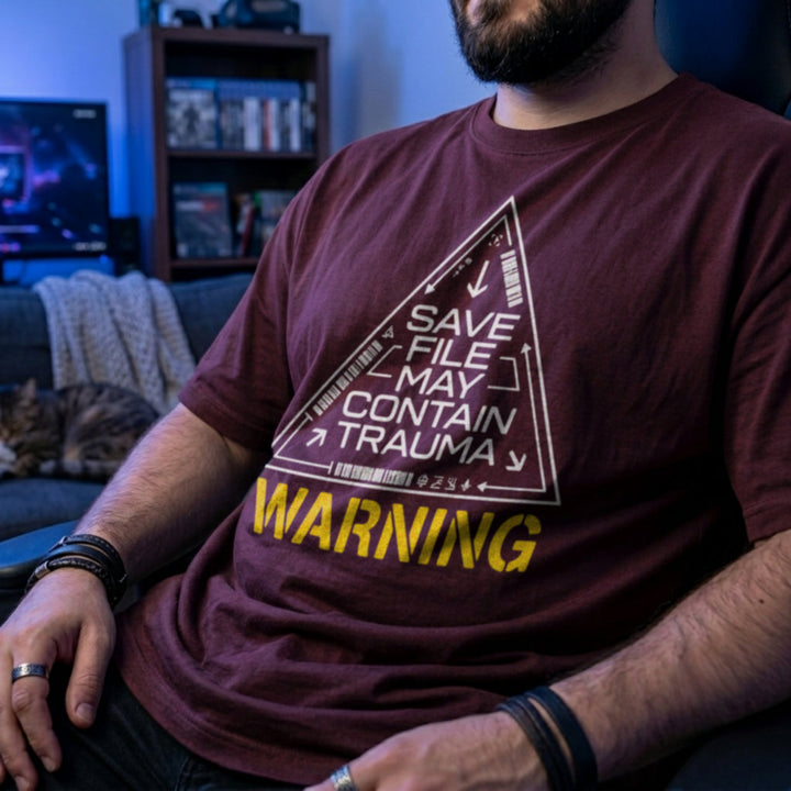 Translation missing: en.Maroon shirt featuring sci-fi triangle warning graphic with text: Save File May Contain Trauma and WARNING label