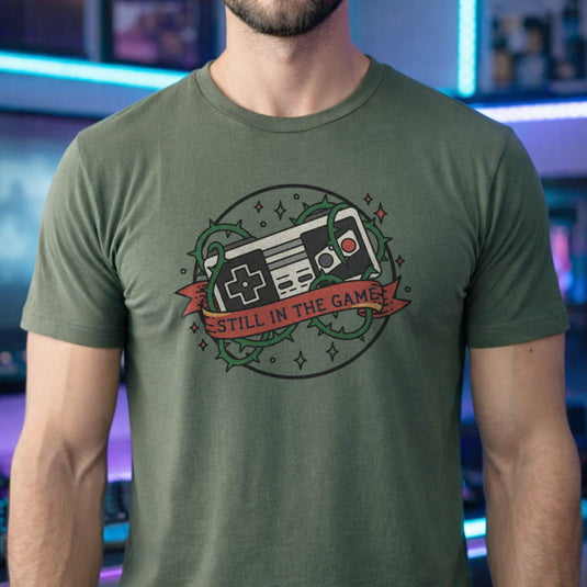 Translation missing: en.Military green  t-shirt with a classic retro controller tangled in green thorns and a banner reading STILL IN THE GAME. 