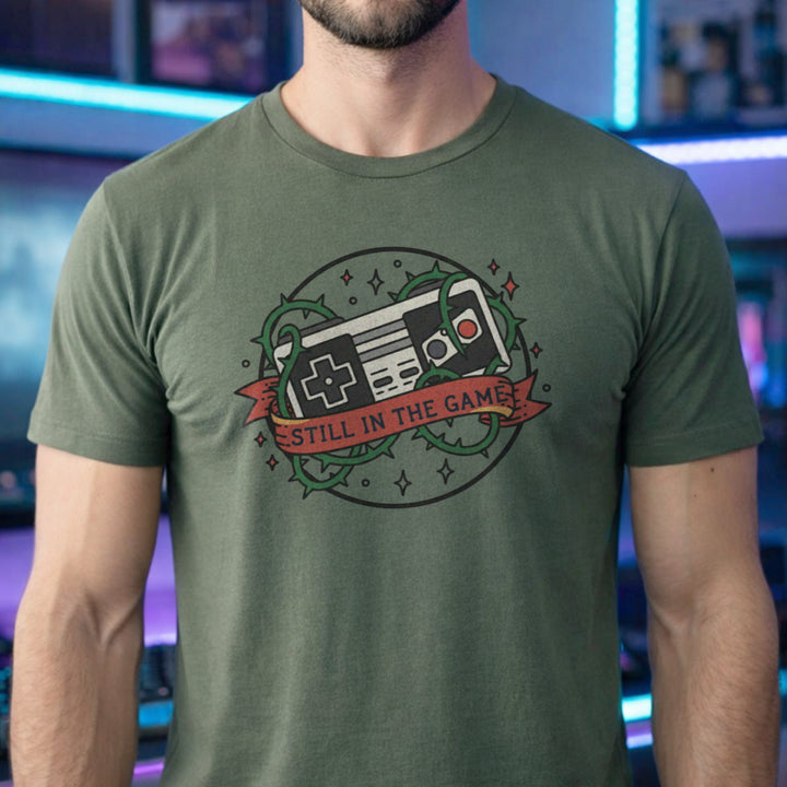 Translation missing: en.Military green  t-shirt with a classic retro controller tangled in green thorns and a banner reading STILL IN THE GAME. 